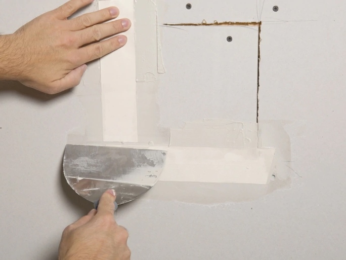 Hands patching a drywall crack with joint compound and mesh tape